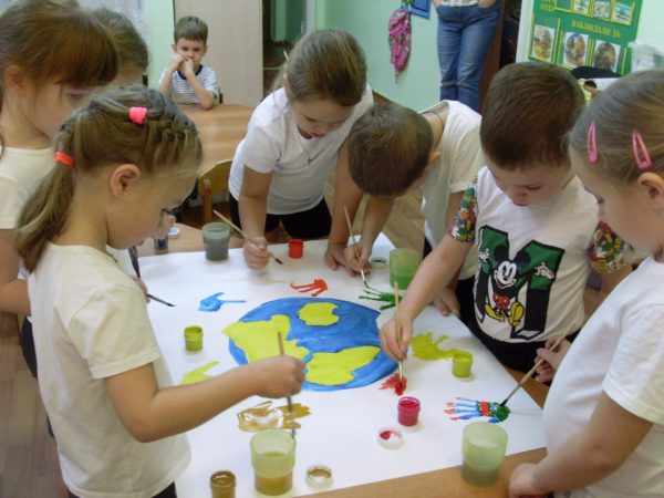 Дети рисуют красками коллективный плакат с изображением земного шара и цветными ладошками