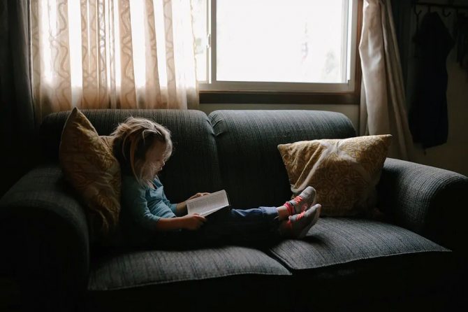 На фото девочка на диване читает книжку.