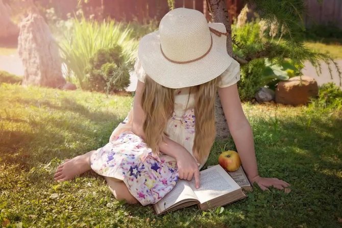 На фото девочка в шляпе читает книжку на траве.